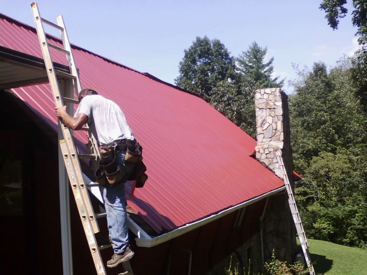 Roofer on a ladder beside a red metal roof and stone chimney — Soffit & Fascia Repair by Roofing Brothers in Stony Point